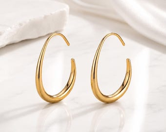 Può includere: Un paio di orecchini a cerchio dorati con un design aperto a forma di goccia. Gli orecchini presentano una finitura liscia e lucida e una silhouette curva e affusolata che crea un effetto ad anello continuo, riflettendo la luce sulla superficie metallica.