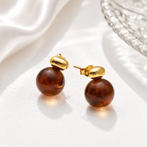 May include: A pair of stud earrings featuring a polished gold-tone metal top piece shaped like a small, smooth oval, connected to a larger, spherical, translucent dark amber-colored bead. The earrings have a post-back closure mechanism.