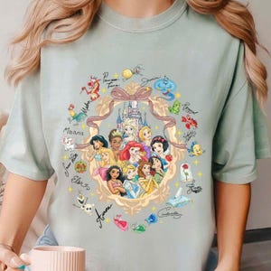 Puede incluir: Camiseta verde claro con un diseño circular que presenta princesas y personajes de Disney. El diseño incluye ilustraciones de princesas, castillos y firmas de personajes. La camiseta tiene cuello redondo y mangas cortas.