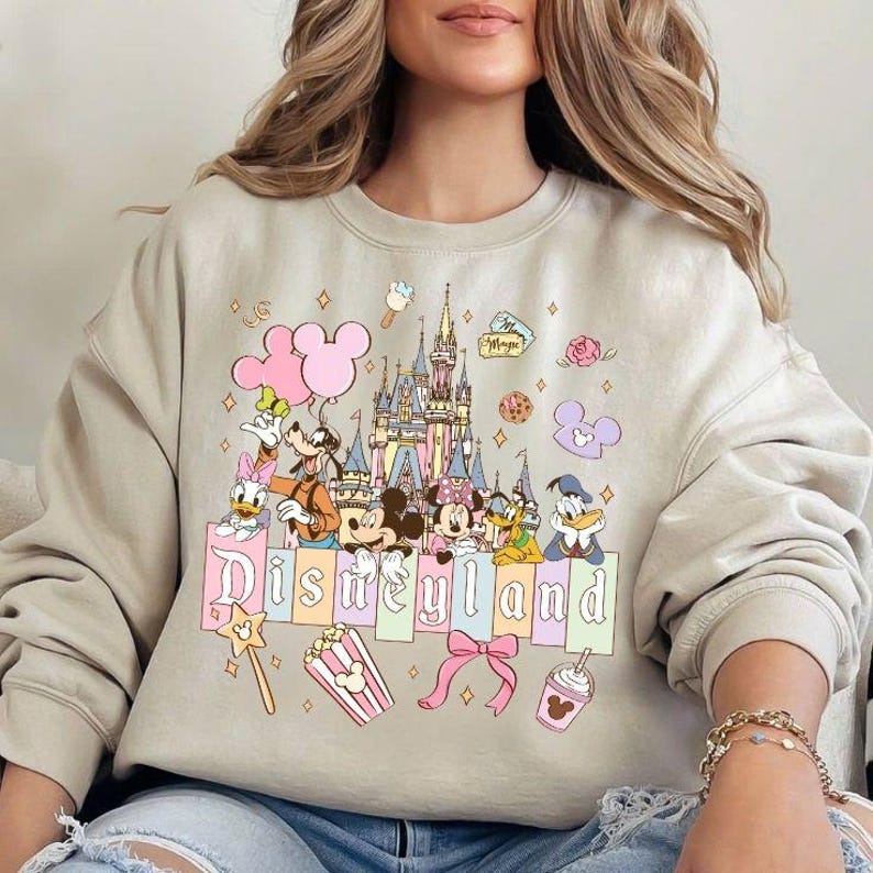 May include: Beige crewneck sweatshirt featuring a colorful Disneyland graphic. The design includes Mickey and Minnie Mouse, Donald Duck, Goofy, and Daisy Duck, with a castle, balloons, and the word "Disneyland". Additional graphics include popcorn, a bow, and a drink.