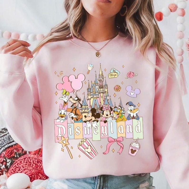 May include: Pink Disneyland sweatshirt featuring Mickey Mouse, Minnie Mouse, Donald Duck, and Goofy. The design includes a castle, popcorn, and the word "Disneyland" in pastel colors. The sweatshirt has a crew neck and long sleeves.