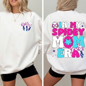 Peut inclure: Sweat-shirt blanc avec un motif de Spider-Gwen. L'avant pr&eacute;sente un graphisme des personnages de Spider-Gwen, tandis que l'arri&egrave;re affiche le texte "IN MY SPIDEY MOM ERA" en rose, bleu et blanc. Le design est lumineux et color&eacute;.