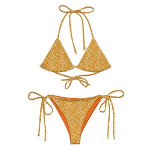 May include: Mustard yellow bikini set with a white floral pattern. The top is a halter style with triangle cups. The bottoms are a tie-side style with an orange lining. The bikini is made of a soft, comfortable material.