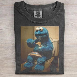 May include: A charcoal gray crew neck t-shirt featuring a graphic print of a blue furry creature sitting on a toilet, eating a cookie with a spoon, and holding a package of cookie dough. The shirt has a Comfort Colors brand label at the neck.