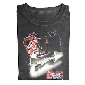 May include: A charcoal gray t-shirt featuring a graphic print of a snarling black panther next to a red Mtn Dew can. The design includes purple lightning bolts and the text Tekken Tag Tournament in a stylized red and white font at the bottom.
