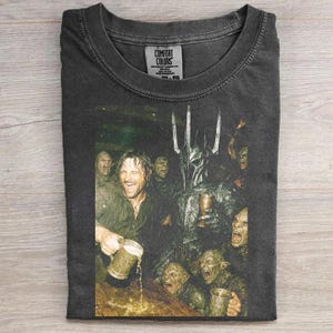 May include: A charcoal gray t-shirt featuring a graphic print of a fantasy scene with a central armored figure in a spiked helmet surrounded by several goblin-like creatures, all holding mugs. The inner neck label reads Comfort Colors.