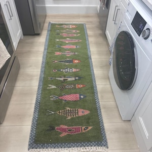May include: A green runner rug with a fish pattern. The rug features a series of stylized fish in various colors, including pink, black, and orange, against a green background. The rug has a blue border and fringe.