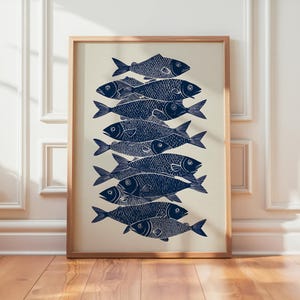 Op de afbeelding: Een ingelijste kunstdruk met een stapel gestileerde marineblauwe vissen op een crèmekleurige achtergrond. De vissen zijn verticaal gerangschikt en overlappen elkaar. Het kunstwerk zit in een houten lijst.