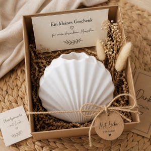 Könnte beinhalten: Eine weiße Muschelschalen-Vase in einer braunen Geschenkbox mit Dekopapier, Trockenblumen und einer kleinen Karte. Auf der Karte steht "Ein kleines Geschenk für einen besonderen Menschen." Ein Anhänger mit Bindfaden sagt "Mit Liebe VERPACKT."