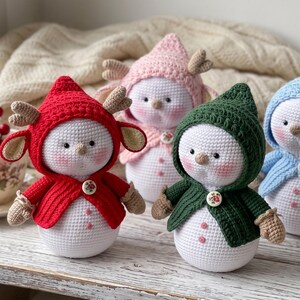 Könnte beinhalten: Vier gehäkelte Schneemannfiguren mit runden weißen Körpern und rosa Wangen. Jede trägt einen Kapuzenumhang in Rot, Rosa, Dunkelgrün oder Hellblau, der mit einem kleinen Blumenknopf befestigt ist. Die roten und rosa Figuren haben kleine Geweihe.