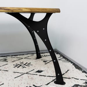 May include: A table with a natural wood grain live edge top and a black metal base. The base features curved, Y-shaped supports secured with visible silver-toned screws, creating a sturdy frame that connects to the underside of the tabletop.
