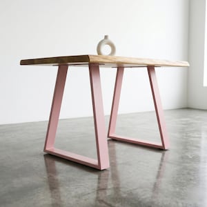 Op de afbeelding: Een tafel met een natuurlijk houten blad met een boomstamrand en twee lichtroze metalen trapeziumvormige poten. Een kleine, witte, cirkelvormige keramische vaas met een gat in het midden staat in het midden op het houten oppervlak.