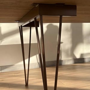 May include: A table with a light brown wooden top and dark brown metal hairpin legs. The legs are attached to a rectangular metal frame mounted underneath the tabletop, providing structural support for the surface above.