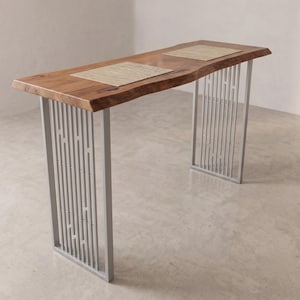 Può includere: Un tavolo console rettangolare con un piano in legno naturale a bordo vivo con venature visibili e due tovagliette intrecciate di colore chiaro. Il tavolo è sostenuto da due gambe in metallo argentato con un design a listelli verticali.