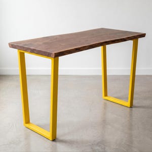 May include: A rectangular table featuring a dark brown natural wood slab top with a live edge and two bright yellow U-shaped metal legs. The table has a smooth, polished surface showing wood grain patterns and a sturdy, modern metal frame construction.