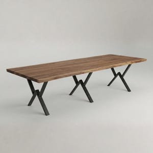 Peut inclure: Une table à manger rectangulaire dotée d'un plateau en bois brun naturel aux bords irréguliers. La table est soutenue par trois pieds en métal noir distincts, chacun construit avec une structure géométrique angulaire en zigzag.
