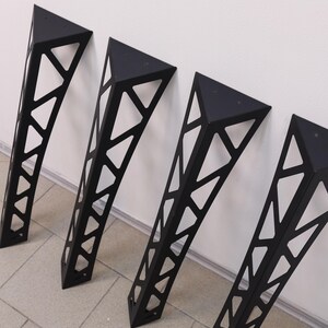 May include: Four matte black metal furniture legs with a triangular cross-section and a geometric open-frame truss design. Each leg features a flat triangular mounting plate at the top with pre-drilled holes for attachment to a flat surface.