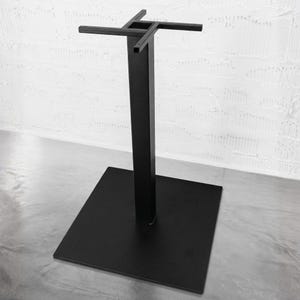May include: A black metal table base featuring a square flat floor plate, a central vertical square column, and a cross-shaped top mounting bracket with four extending arms designed to support a tabletop surface in a stable upright position.