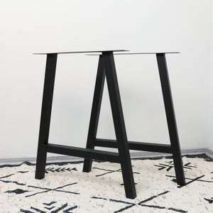 Può includere: Una coppia di gambe per tavolo in metallo nero con design a telaio ad A. Ogni struttura è composta da due supporti verticali inclinati collegati da una traversa orizzontale alla base e da una piastra di montaggio piatta sulla parte superiore.