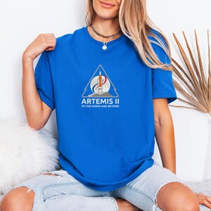 May include: A royal blue t-shirt featuring the Artemis II mission logo. The logo includes a rocket, a crescent moon, and the text "ARTEMIS II TO THE MOON AND BEYOND" in white. The shirt is a crew neck style.