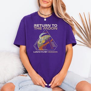 May include: A purple t-shirt with the text "RETURN TO THE MOON" in gold lettering. Below the text is a graphic of a moon and a rocket, with the words "LUNAR FLYBY MISSION". The shirt is a short-sleeved, crew neck style.