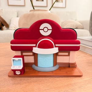 May include: A red and white 3D-printed model of a building with a circular logo on top. The building has a blue door and a small red machine to the left. The model sits on a wooden surface.