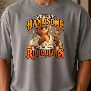 May include: A grey short-sleeve t-shirt featuring a graphic of a brown bear wearing sunglasses and a white bathrobe while holding a hand mirror. The text reads WOKE UP HANDSOME AGAIN THIS IS GETTING RIDICULOUS in orange and yellow lettering.