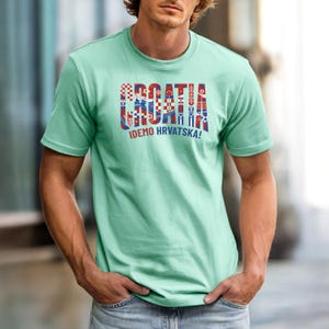 May include: A light green t-shirt with the word "CROATIA" in a colorful, patterned design. Below the word is the phrase "IDEMO HRVATSKA!" in white. The shirt is short-sleeved.
