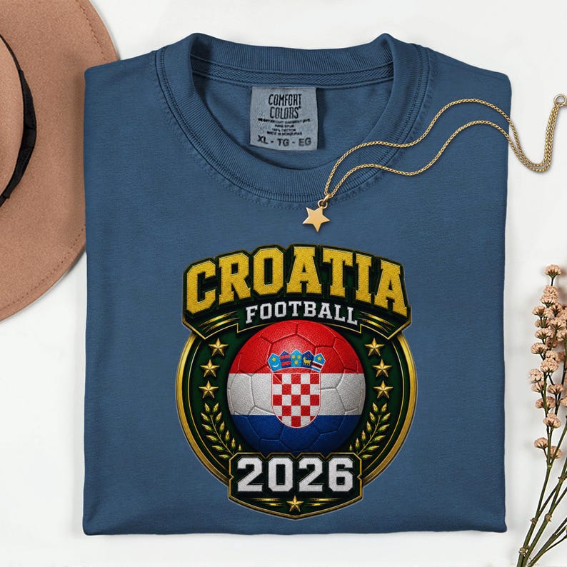 Peut inclure: Un t-shirt bleu avec un motif de football croate. Le motif comprend un ballon de football avec le drapeau croate, le mot "CROATIA" en or et l'ann&eacute;e "2026". Un collier &eacute;toile dor&eacute; est drap&eacute; sur le t-shirt.