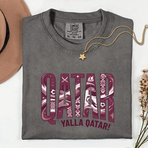 Puede incluir: Una camiseta gris Comfort Colors con la palabra "QATAR" en granate y blanco, con patrones decorativos y el texto "YALLA QATAR!". Un collar con colgante de estrella dorada descansa sobre la camiseta.