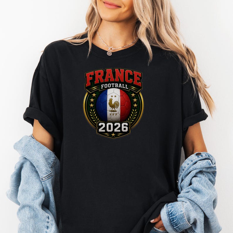 以下が含まれることがあります： フランスサッカーのグラフィックデザインが施された黒いTシャツ。デザインには、赤い文字で「FRANCE FOOTBALL」、フランス国旗の色を使ったサッカーボール、そして年号「2026」が含まれています。デザインは金の月桂樹の冠で縁取られています。