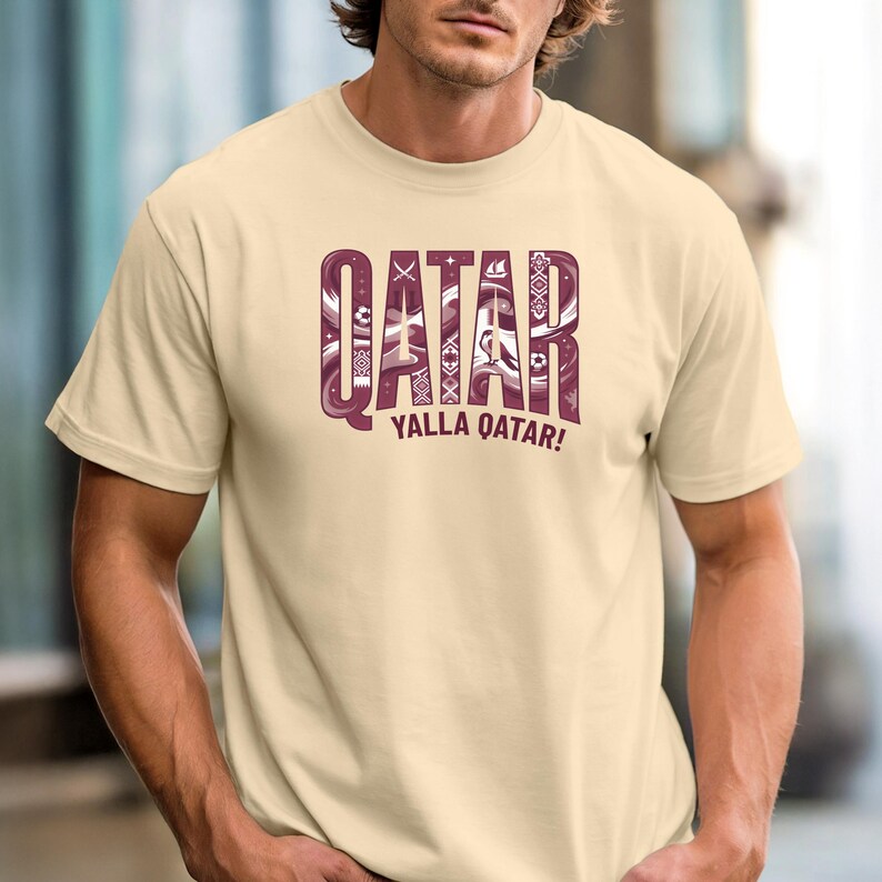 Puede incluir: Una camiseta beige con la palabra "QATAR" en una fuente decorativa de color granate. El texto "YALLA QATAR!" est&aacute; debajo. La camiseta es de manga corta.