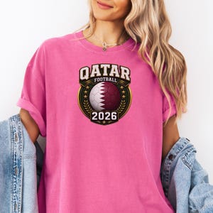 May include: A pink t-shirt featuring a Qatar Football design. The design includes a soccer ball with the Qatar flag colors, the text "QATAR FOOTBALL" and the year "2026" in a gold and green emblem.