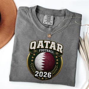 May include: Gray t-shirt with a Qatar Football graphic. The graphic features a soccer ball in the colors of the Qatar flag, the text "QATAR FOOTBALL" and the year "2026". A silver necklace with a pendant is on the shirt.
