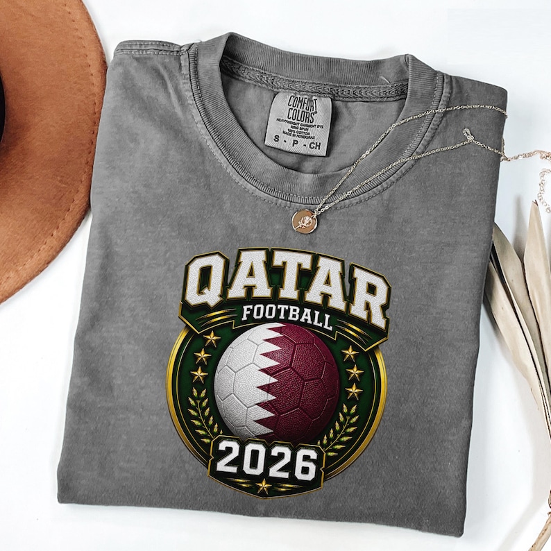 May include: Gray t-shirt with a Qatar Football graphic. The graphic features a soccer ball in the colors of the Qatar flag, the text "QATAR FOOTBALL" and the year "2026". A silver necklace with a pendant is on the shirt.