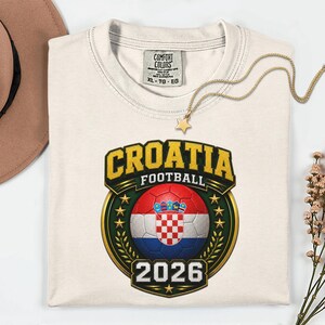 Peut inclure: T-shirt cr&egrave;me avec un motif de football croate. Le motif comprend le mot "CROATIA" en lettres dor&eacute;es, un ballon de football avec le drapeau croate et l'ann&eacute;e "2026". Un collier dor&eacute; avec un pendentif en forme d'&eacute;toile est pos&eacute; sur le t-shirt.