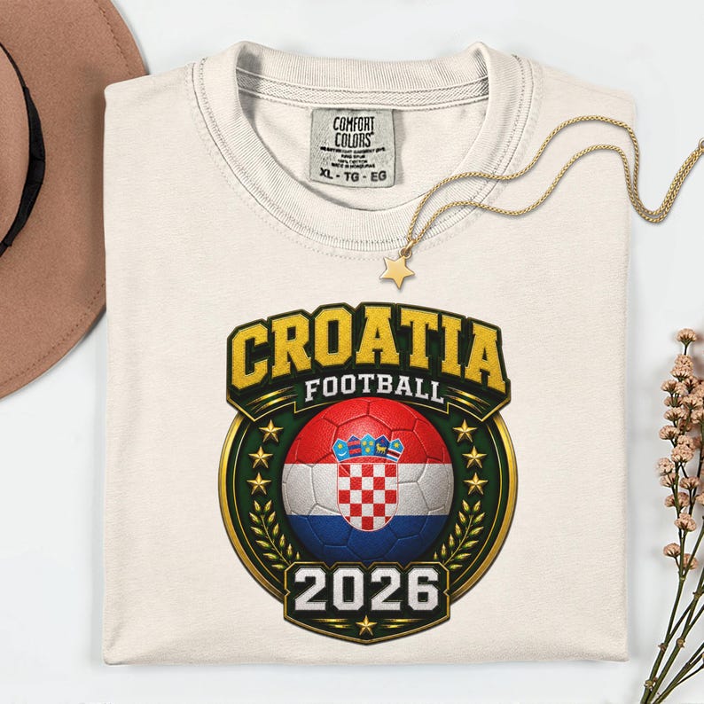 Peut inclure: T-shirt cr&egrave;me avec un motif de football croate. Le motif comprend le mot "CROATIA" en lettres dor&eacute;es, un ballon de football avec le drapeau croate et l'ann&eacute;e "2026". Un collier dor&eacute; avec un pendentif en forme d'&eacute;toile est pos&eacute; sur le t-shirt.