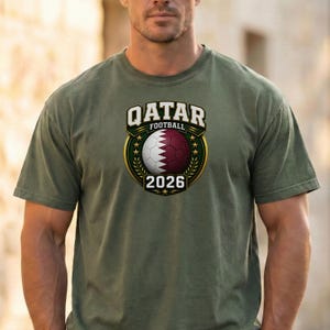 May include: Olive green t-shirt featuring a Qatar Football emblem. The emblem includes a soccer ball in maroon and white, the text "QATAR FOOTBALL" above, and the year "2026" below. The design is framed by a gold border.