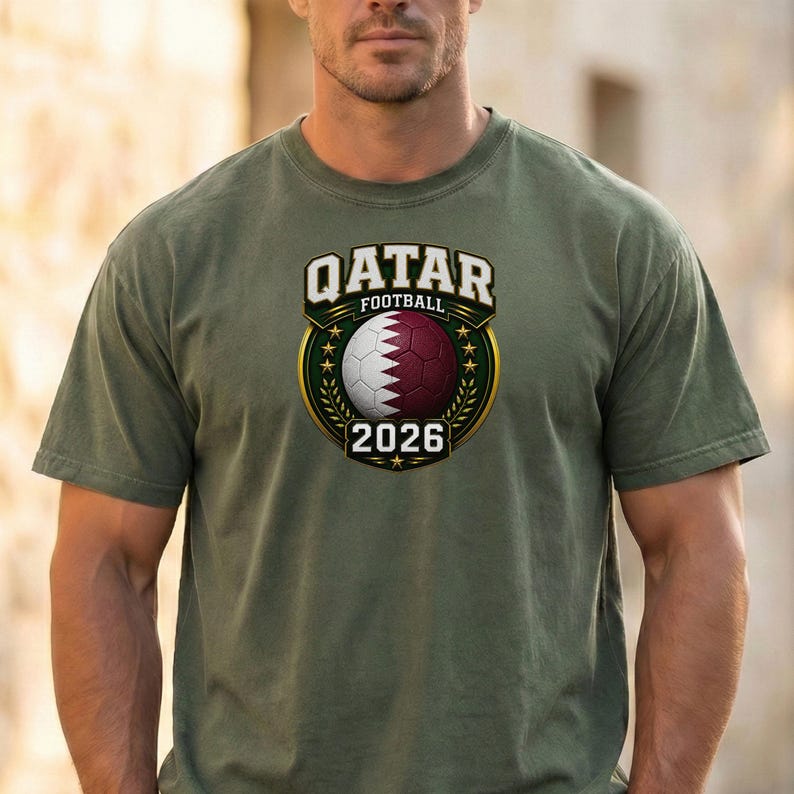 May include: Olive green t-shirt featuring a Qatar Football emblem. The emblem includes a soccer ball in maroon and white, the text "QATAR FOOTBALL" above, and the year "2026" below. The design is framed by a gold border.
