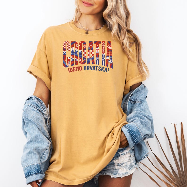 May include: Mustard yellow t-shirt with the word "CROATIA" in red, white, and blue block letters. The text "IDEMO HRVATSKA!" is printed below the word "CROATIA". The shirt is worn with a denim jacket and shorts.