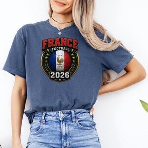 以下が含まれることがあります： フランスの国旗の色、雄鶏、そして「FRANCE FOOTBALL 2026」の文字が入ったサッカーボールのグラフィックが特徴の青いTシャツ。 デザインは金と緑のエンブレムの中にあります。
