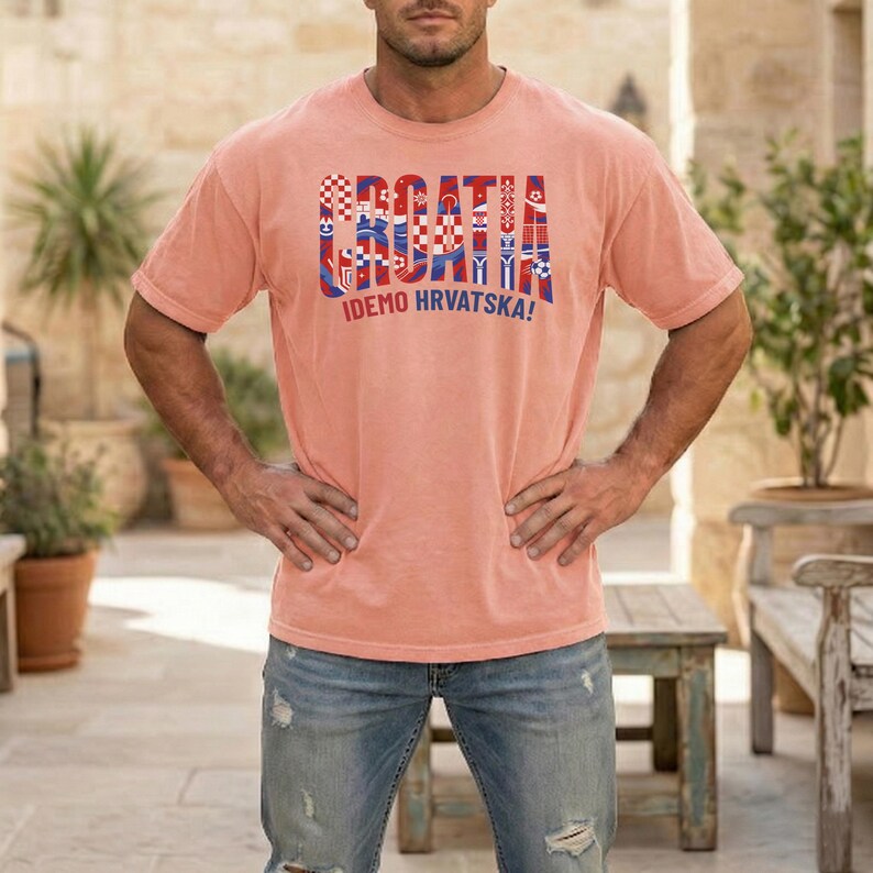 May include: A coral-colored t-shirt with the word "CROATIA" in a multi-colored, patterned design. Below the word is the phrase "IDEMO HRVATSKA!" in blue. The shirt is short-sleeved and has a relaxed fit.