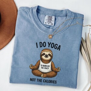 May include: A light blue t-shirt with the text "I DO YOGA" and a cartoon sloth doing yoga. The sloth wears a headband and a shirt that says "TO BURN OFF THE CRAZY NOT THE CALORIES". A gold necklace is draped across the shirt.