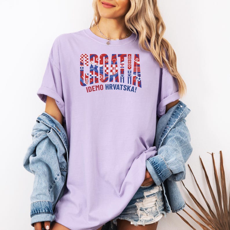 May include: A light purple t-shirt with the word "CROATIA" in a red, white, and blue design. Below the word "CROATIA" is the text "IDEMO HRVATSKA!" The shirt is a casual style.
