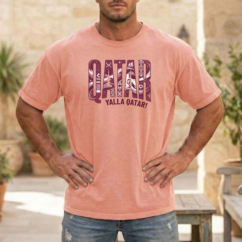 Puede incluir: Una camiseta de color coral con la palabra "QATAR" en una fuente decorativa y estampada. Debajo de la palabra est&aacute; la frase "YALLA QATAR!". La camiseta tiene mangas cortas y la lleva una persona.