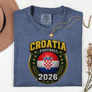 Peut inclure: Un t-shirt bleu avec un graphisme repr&eacute;sentant le mot "CROATIA" en lettres dor&eacute;es au-dessus d'un ballon de football avec le drapeau croate. Le texte "FOOTBALL" est en dessous du mot "CROATIA", et l'ann&eacute;e "2026" est en bas.