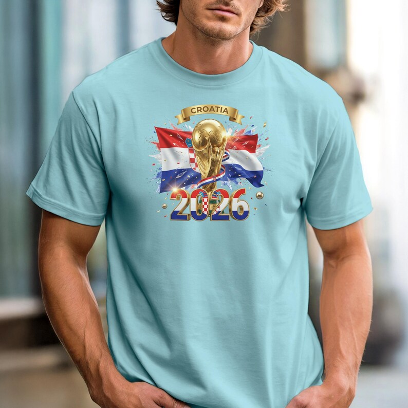 May include: Light blue t-shirt featuring a graphic design with the Croatian flag, a golden trophy, and the text "Croatia 2026". The design includes red, white, and blue colors, with gold accents. The shirt has short sleeves and a crew neck.