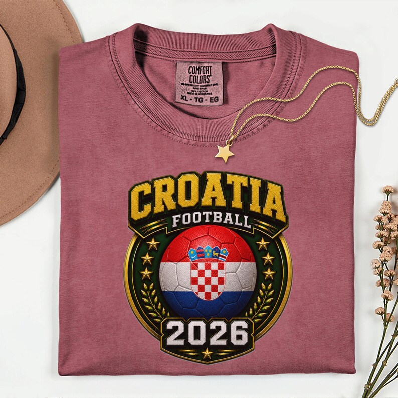 Peut inclure: Un t-shirt rose poussi&eacute;reux avec un graphisme Croatie Football. Le graphisme pr&eacute;sente un ballon de football aux couleurs du drapeau croate, le mot "CROATIA" en or et l'ann&eacute;e "2026". Un collier pendentif &eacute;toile en or est drap&eacute; sur le t-shirt.