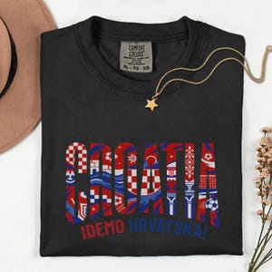 May include: Black Comfort Colors t-shirt with the word "CROATIA" in red, white, and blue, featuring various Croatian-themed designs. The text "IDEMO HRVATSKA!" is below. A gold star pendant necklace and a brown hat are also visible.