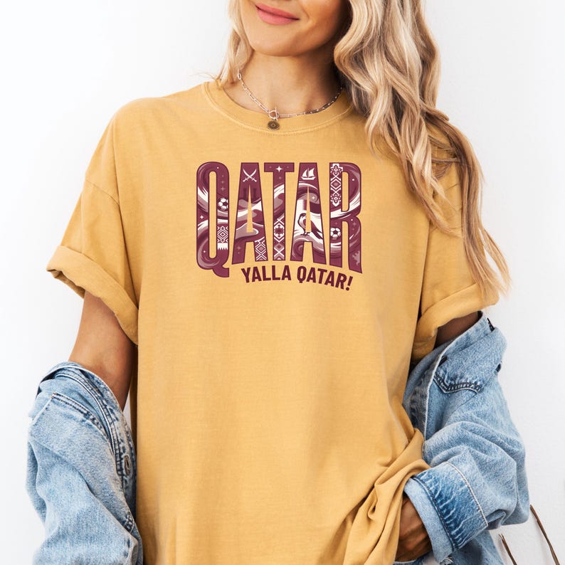 Puede incluir: Camiseta de color amarillo mostaza con la palabra "QATAR" en un dise&ntilde;o granate estampado. Debajo, la frase "YALLA QATAR!" en granate. La camiseta tiene las mangas dobladas y se lleva con una chaqueta vaquera.